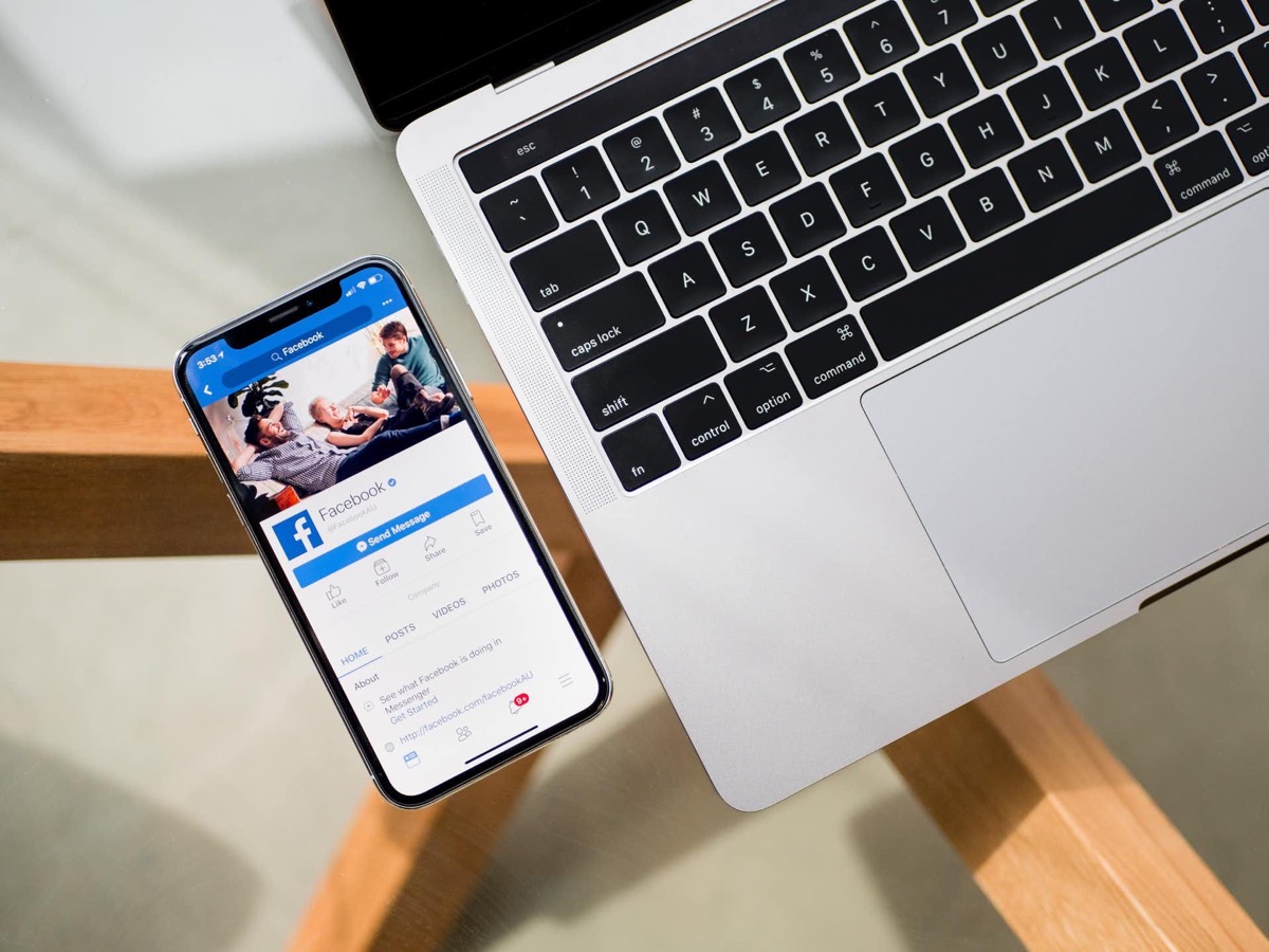 Ein Smartphone mit geöffneter Facebook-App und ein Laptop liegen auf einem Holztisch.